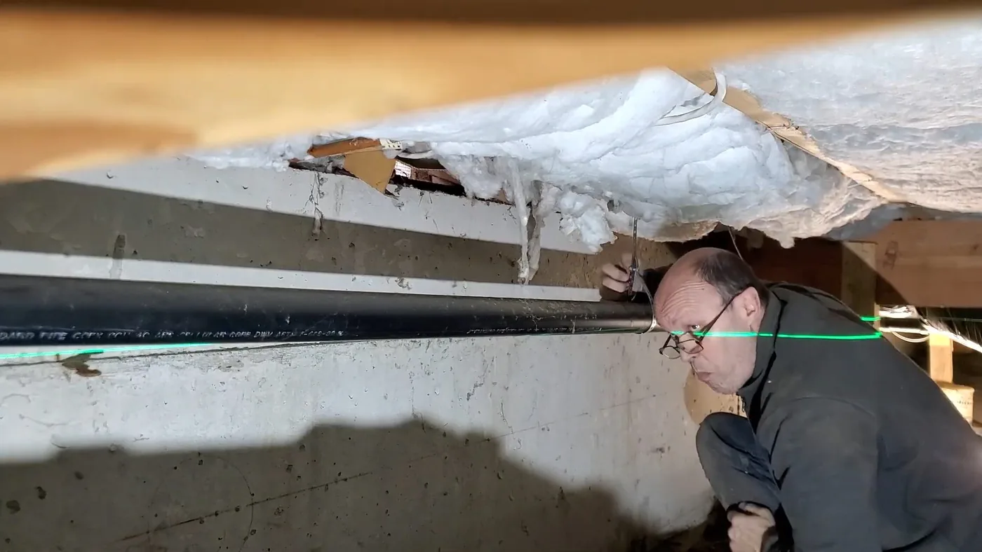 Man crouching in a crawl space beside a black pipe, with torn fiberglass insulation hanging from the floor above