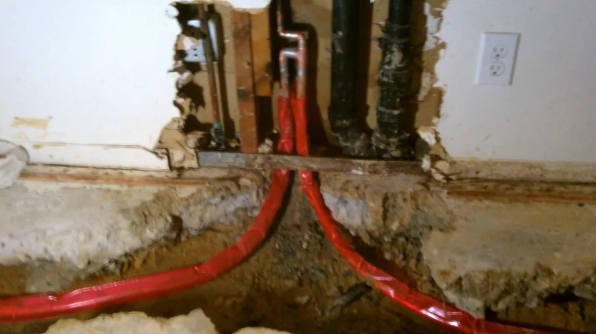 Cut open wall base with red and black pipes passing through damp looking debris and insulation