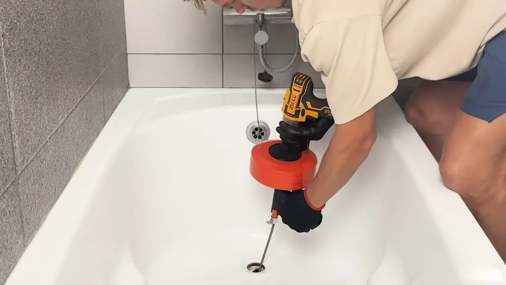 Man leaning over a bathtub, feeding a drum drain snake into the drain with a cordless drill