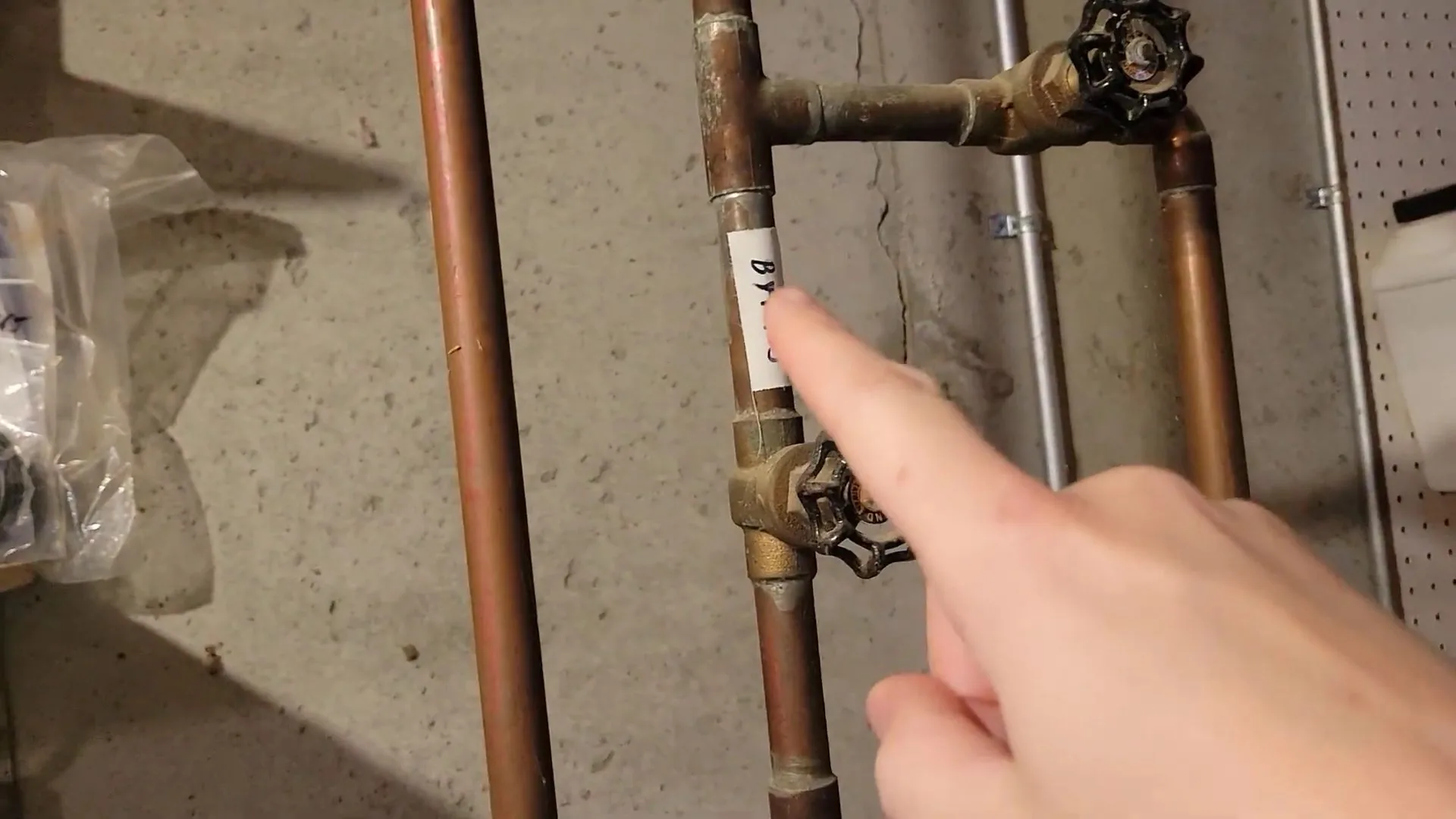 Finger pointing at a pair of brass shutoff valves on copper pipes in front of a concrete wall