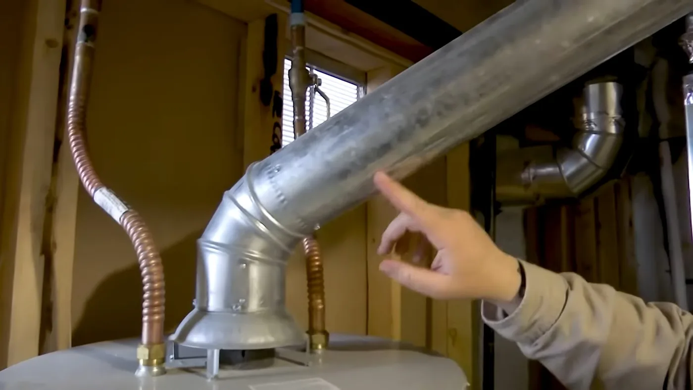 Finger pointing at the metal vent connection on top of a water heater, with copper and flexible gas lines beside it