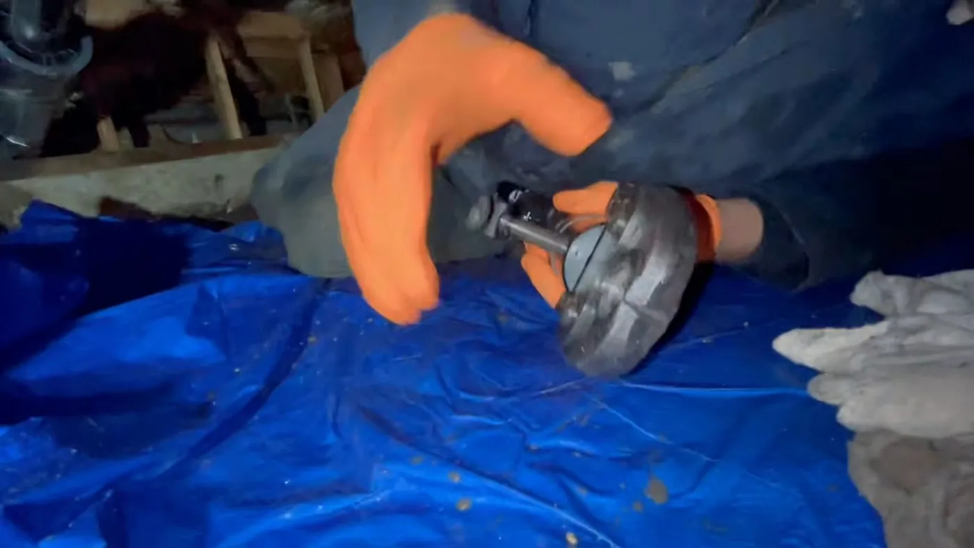 Gloved hands holding a metal fitting over a blue tarp in a crawl space
