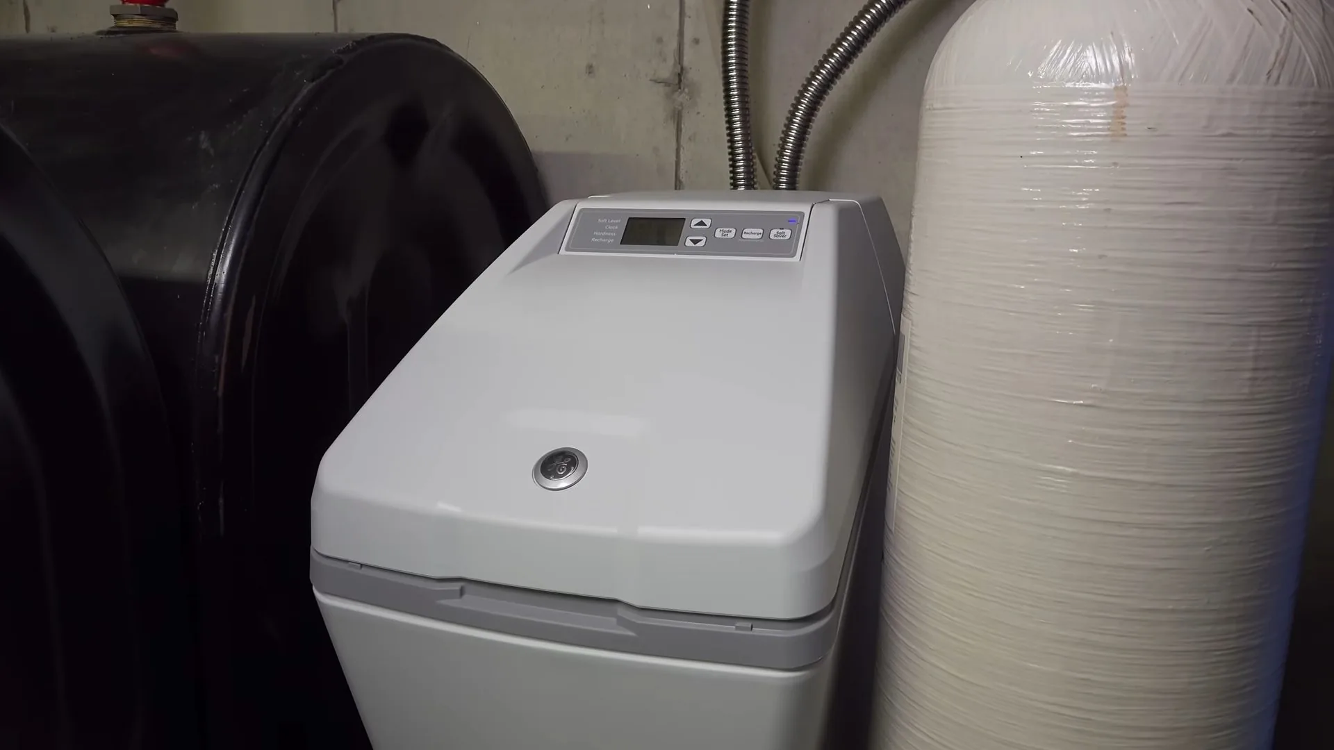 Compact gray water softener standing between a black tank and a tall white tank in a utility corner