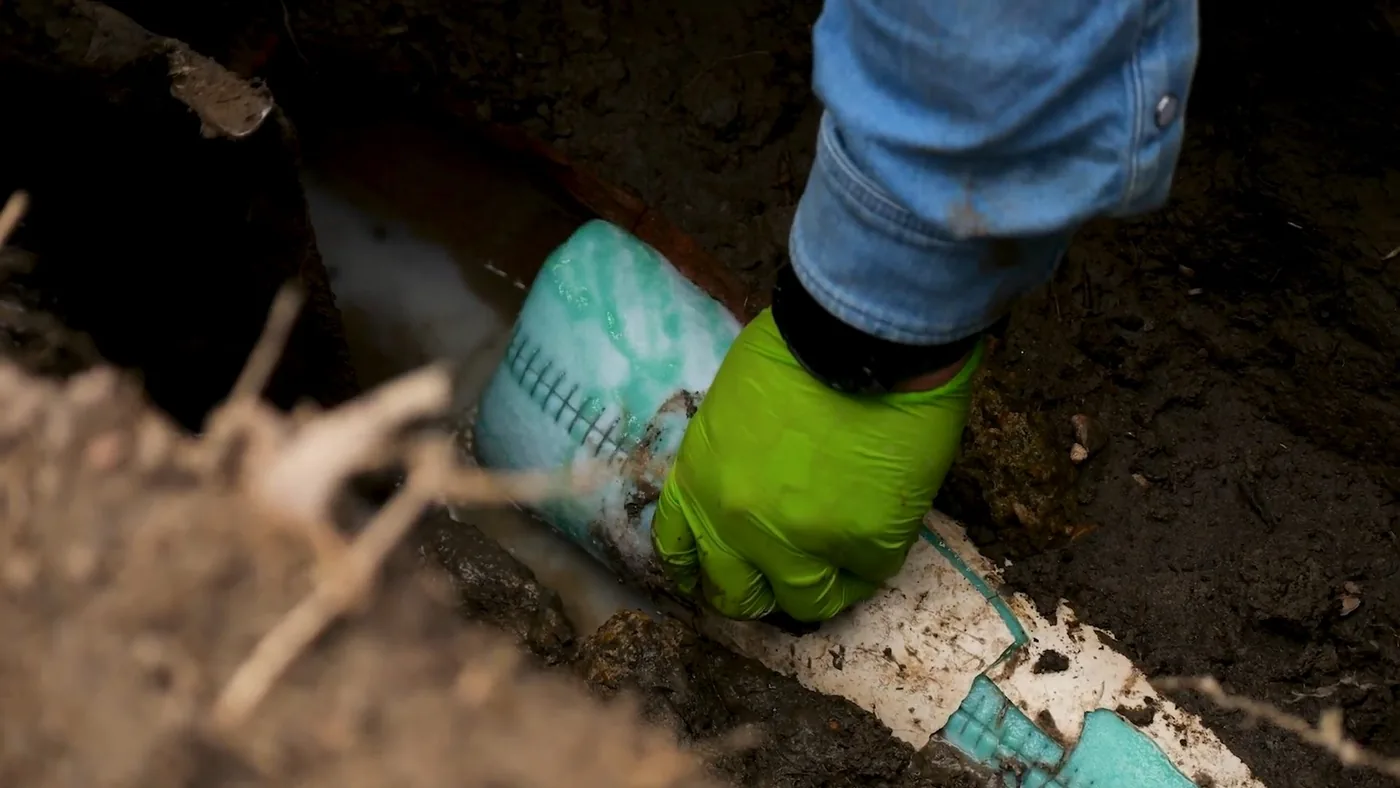 Green-gloved hand setting a liner section into a small dirt pit beside a white pipe