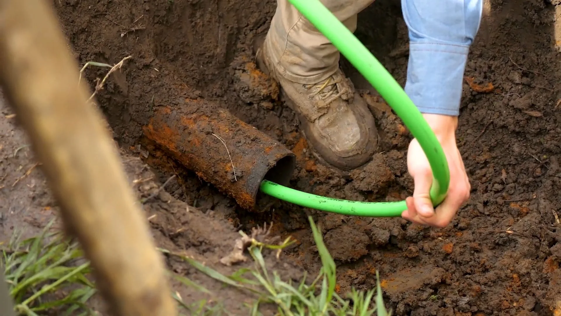 Man in work boots guiding a green hose into the open end of a rusty pipe in a dirt trench