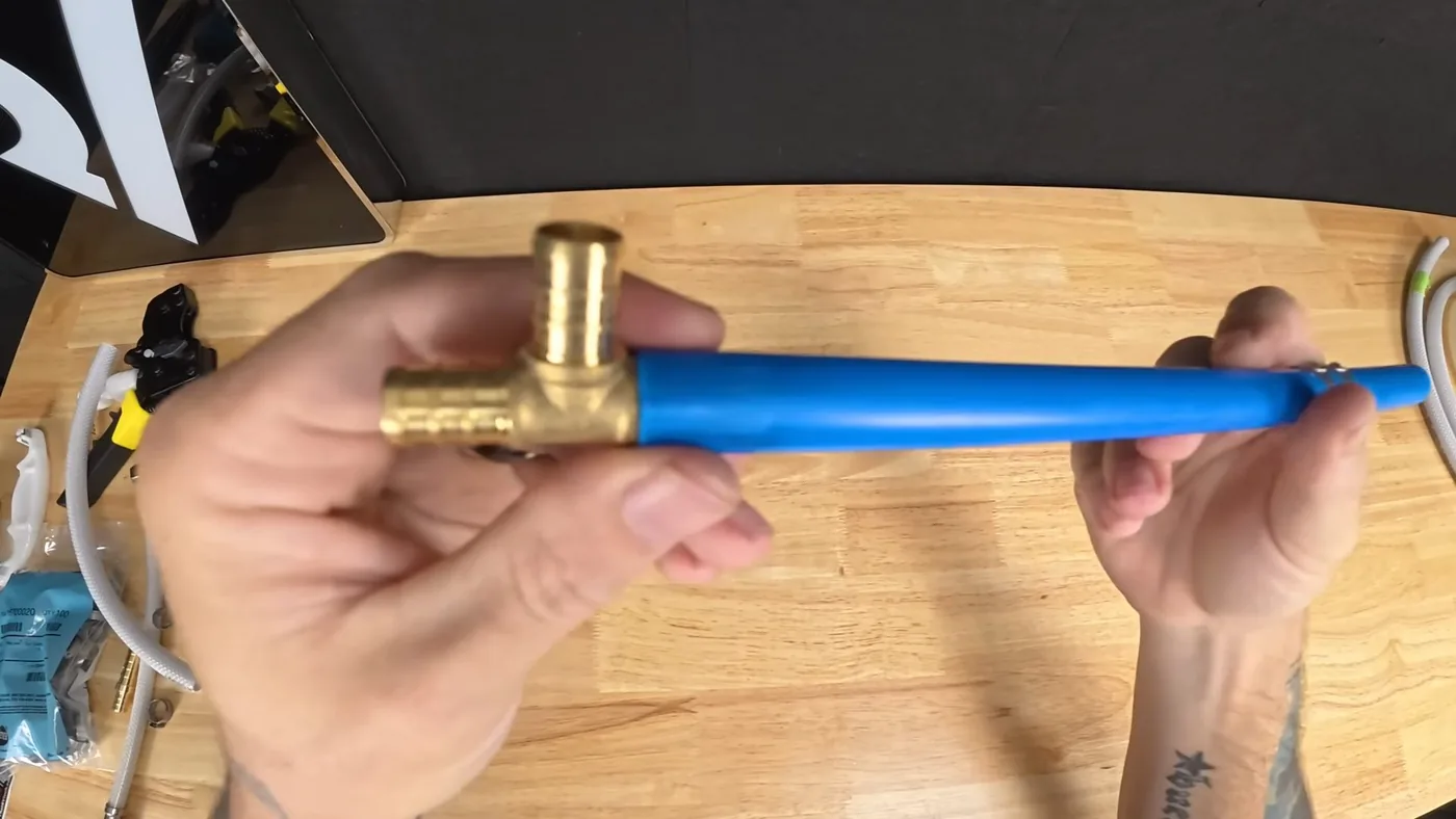 Hand holding a length of blue PEX tubing fitted with a brass tee over a wooden bench