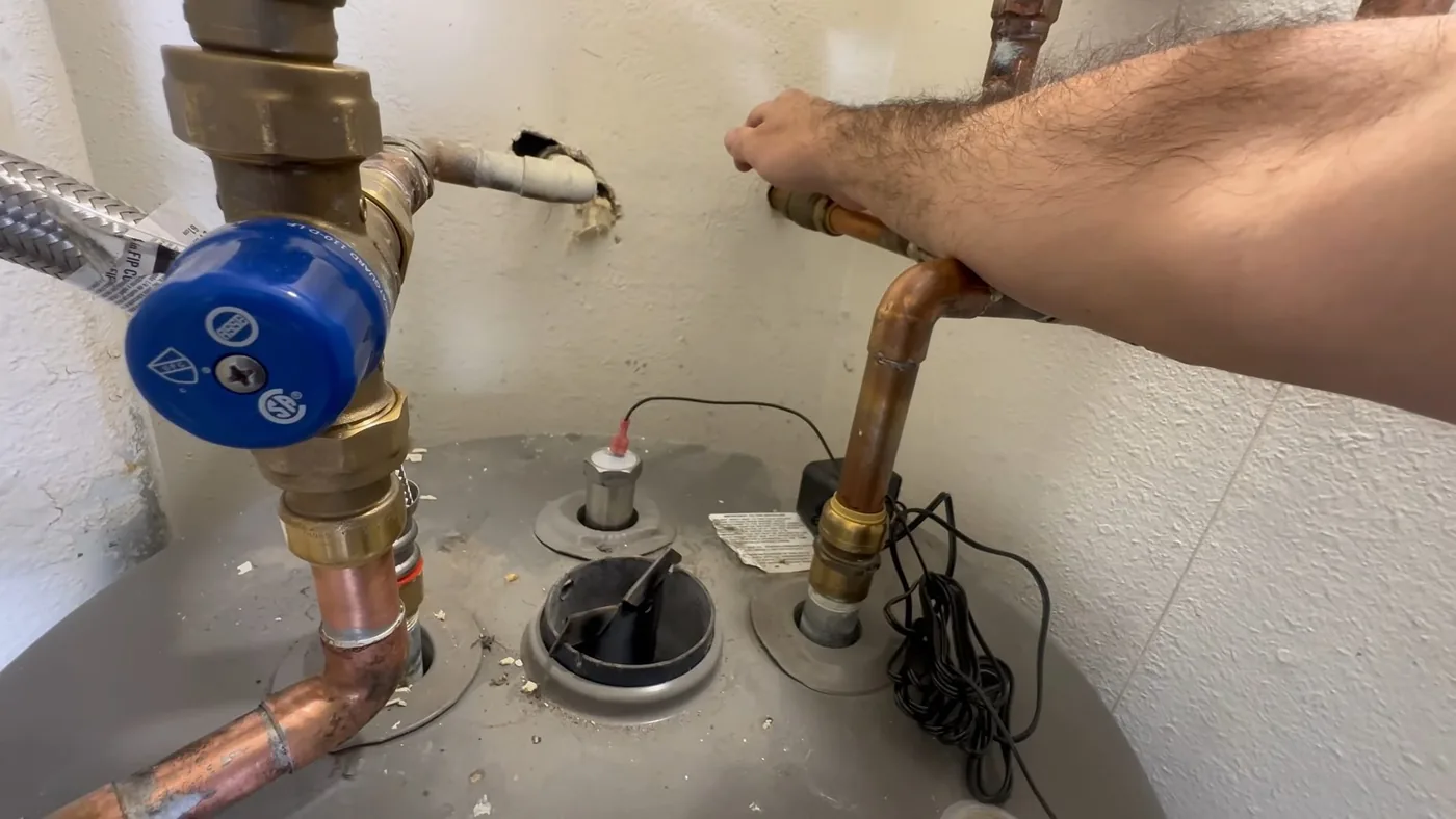 Hand holding a copper line on top of a gray tank water heater beside two valves and a flexible connector