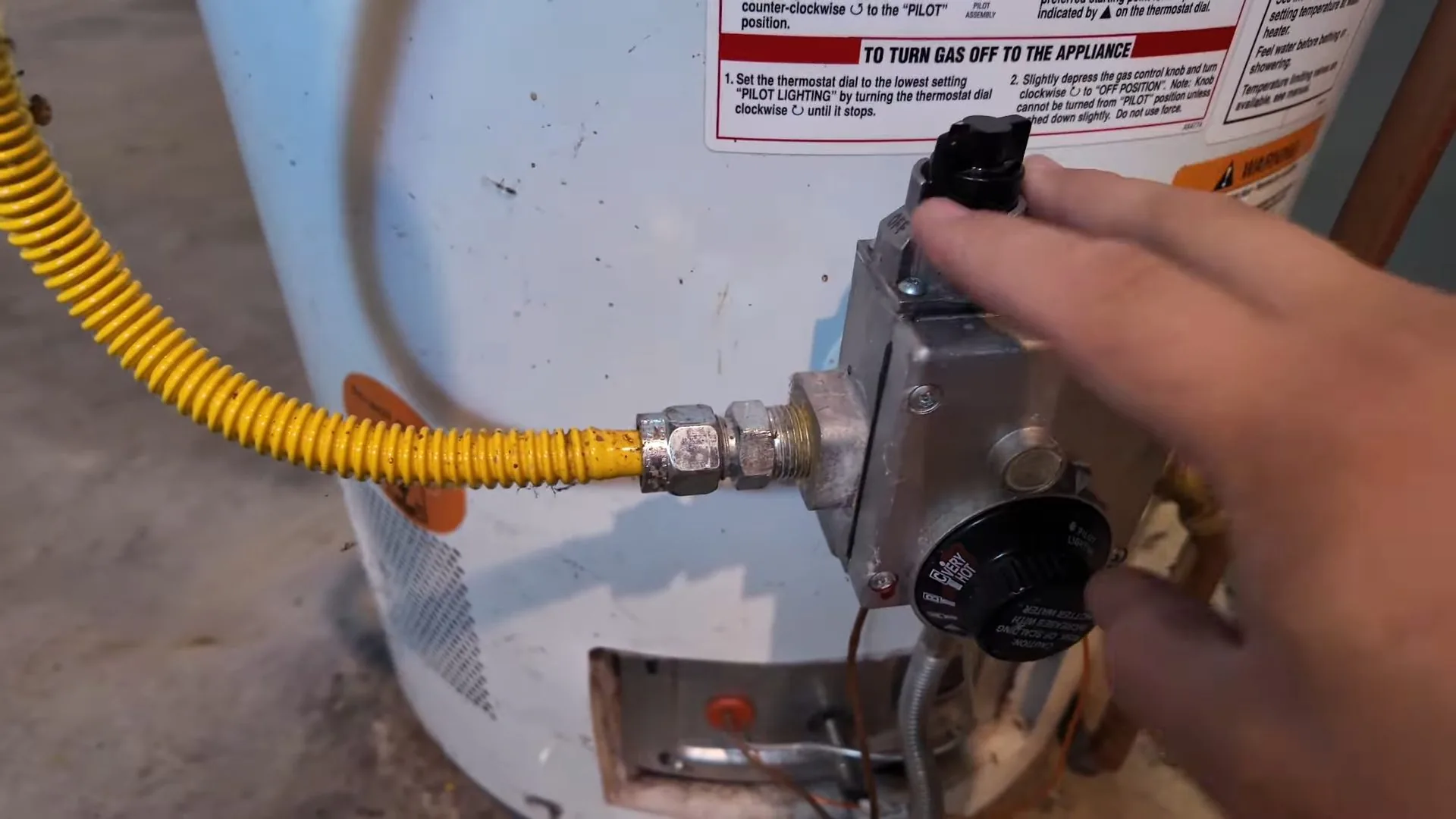 Hand on the knob of a water heater gas control, with a yellow flexible gas line running to it