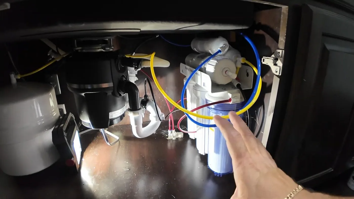 Hand reaching under a sink toward a garbage disposal and a set of water filter housings with colored tubing