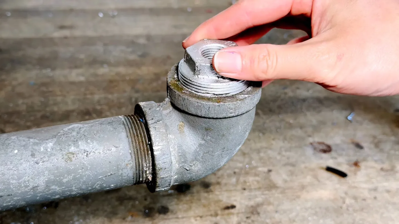 Hand turning a threaded plug out of a galvanized pipe elbow lying on a worktable