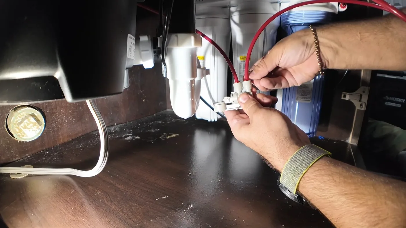 Hands joining two small fittings on colored tubing beside a garbage disposal under a sink
