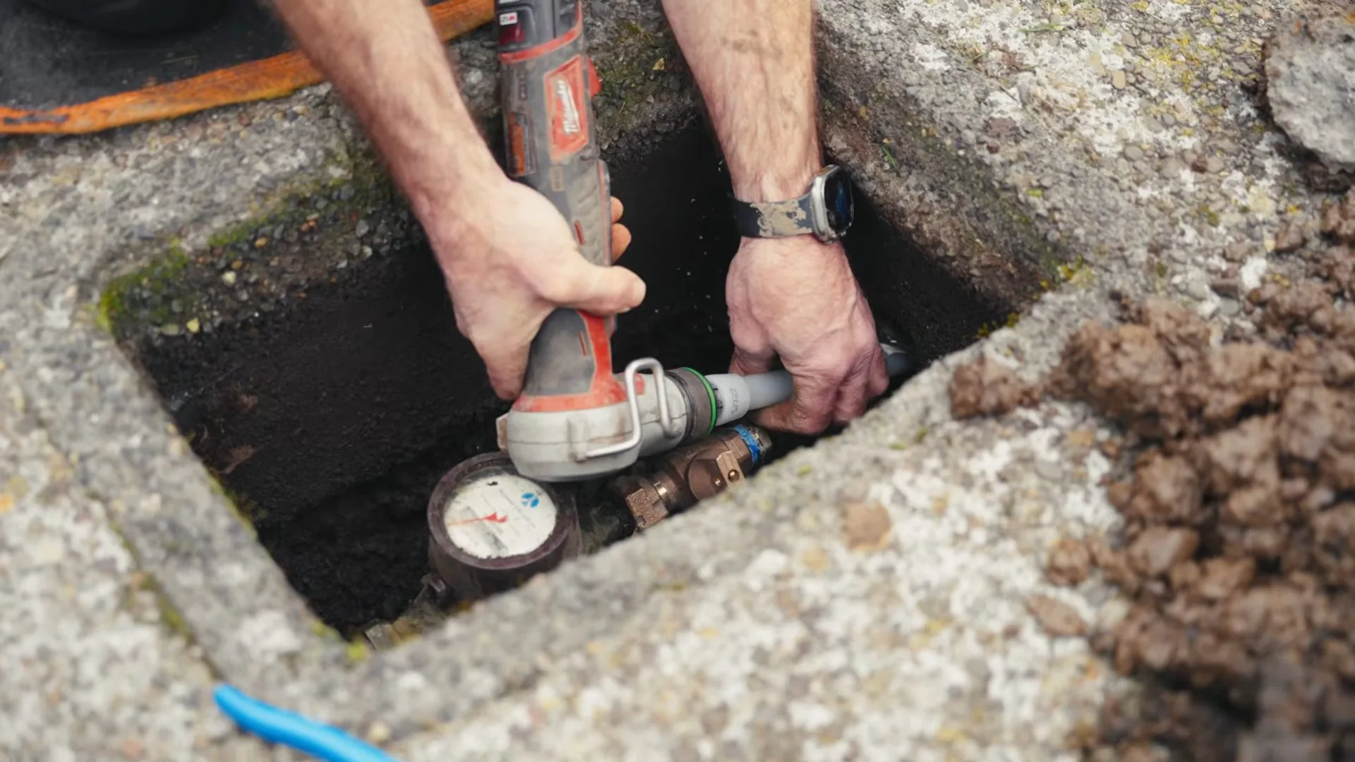 Hands holding a power tool inside a concrete meter box, with a water meter dial and a valve at the bottom