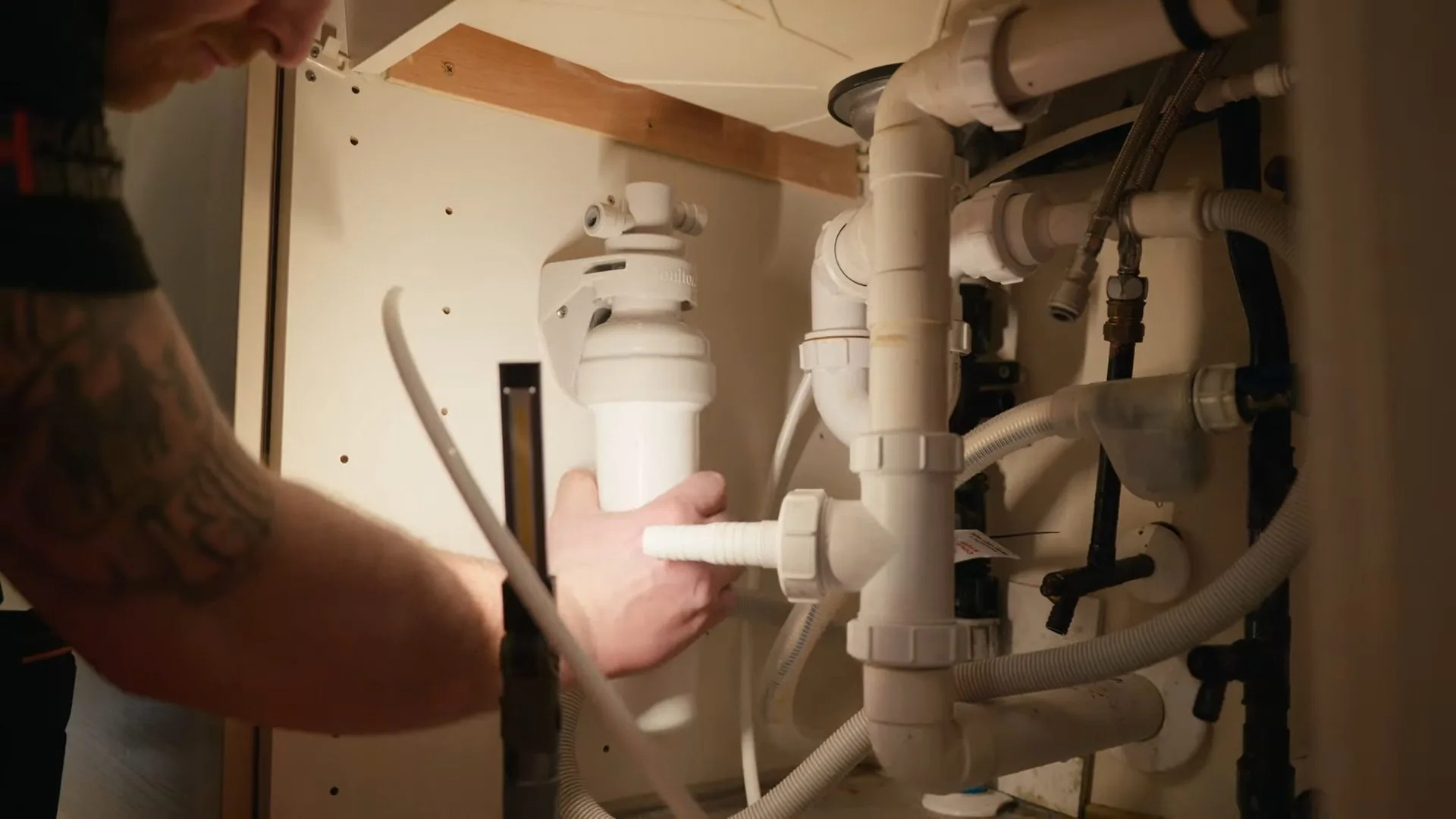 Hands working on a cluster of white drain and supply pipes inside a sink cabinet