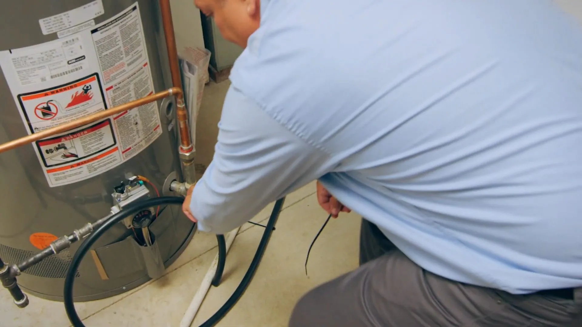 Man in a light blue shirt crouching beside a tank water heater with a dark hose running to its drain