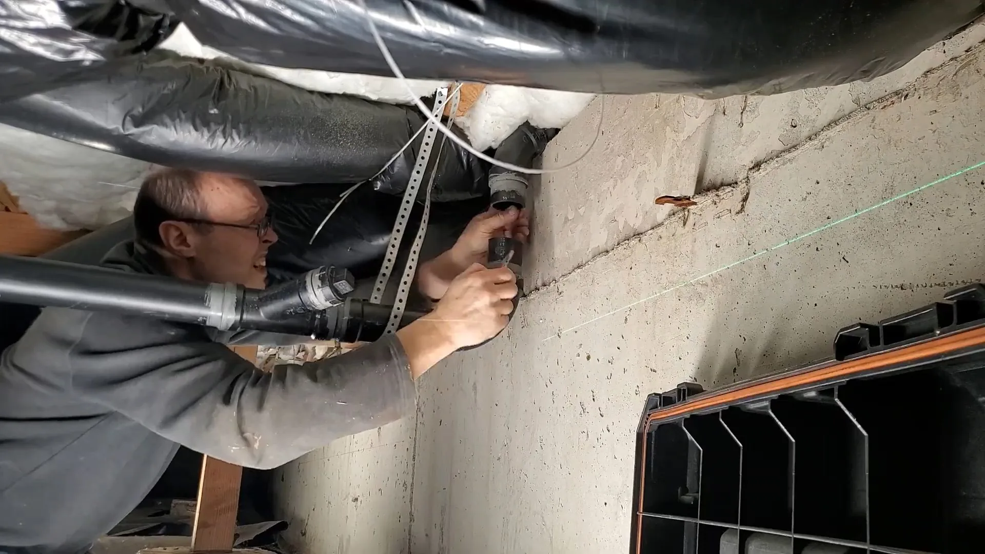 Man in a crawl space beside a concrete foundation wall, reaching toward a pipe near the floor above