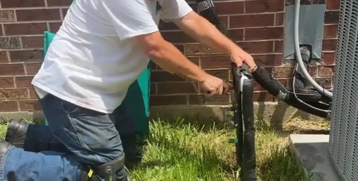Man kneeling on grass beside a black pipe assembly against a brick wall next to an outdoor unit