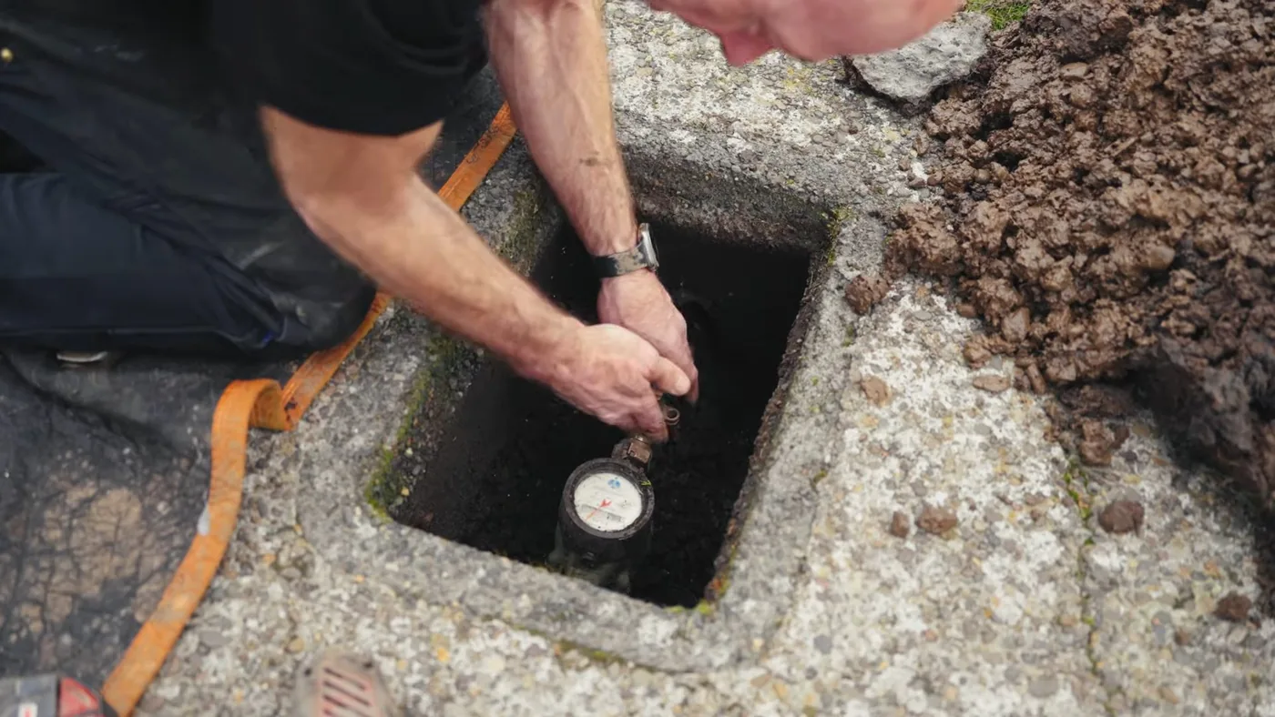 Man kneeling beside an open concrete meter box with one hand on the water meter inside