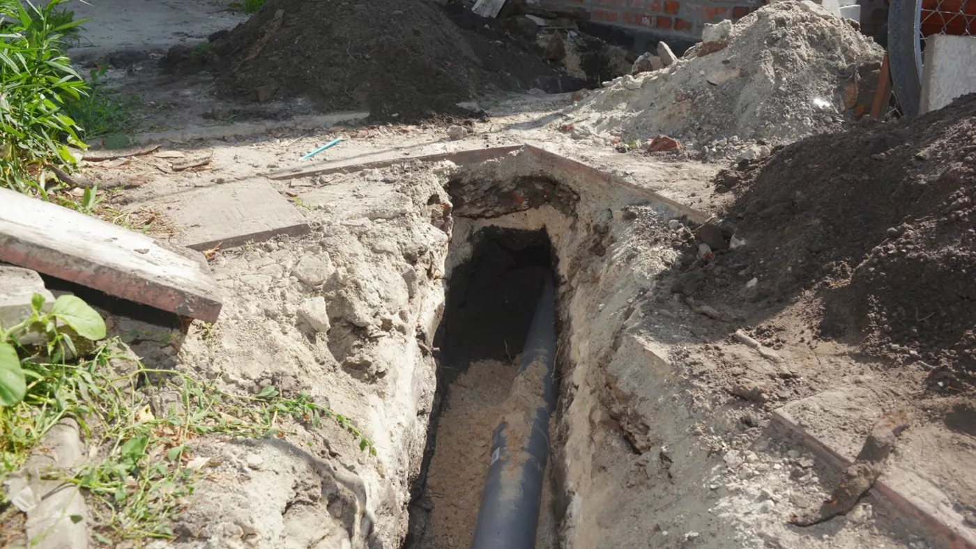 Narrow trench in a yard with a dark pipe visible at the bottom and loose soil piled beside it