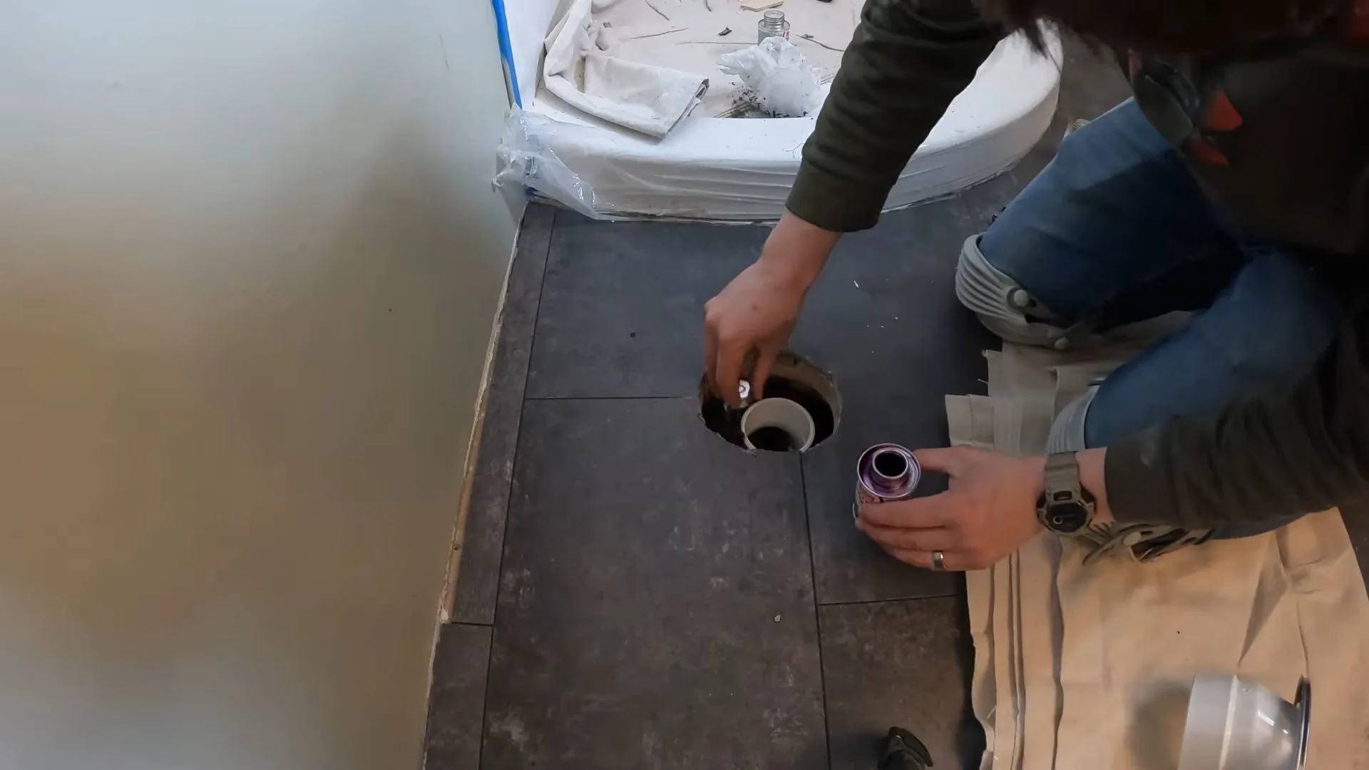 Person kneeling beside an open toilet drain flange on a dark floor, with cleaning supplies nearby