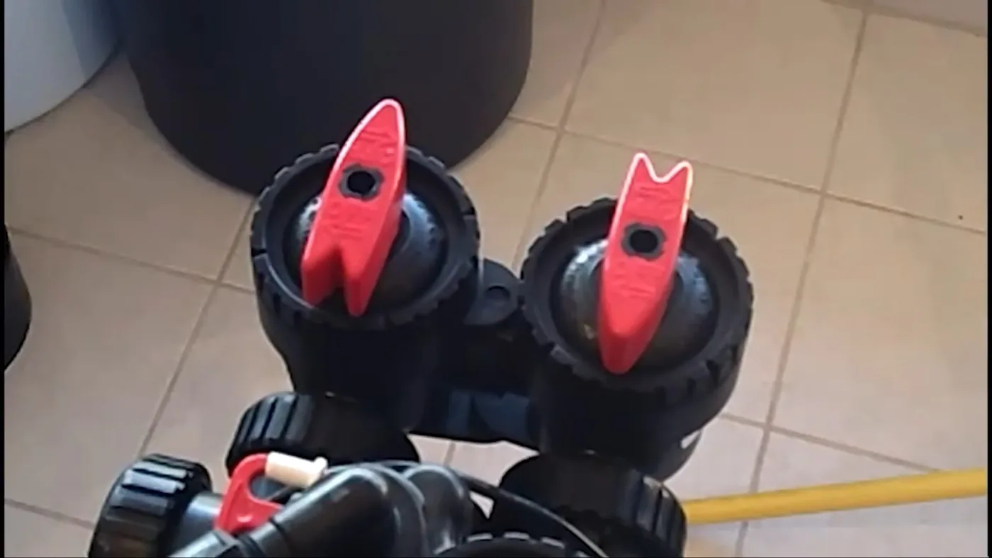 Two red valve handles on the head of a water softener above a tiled floor