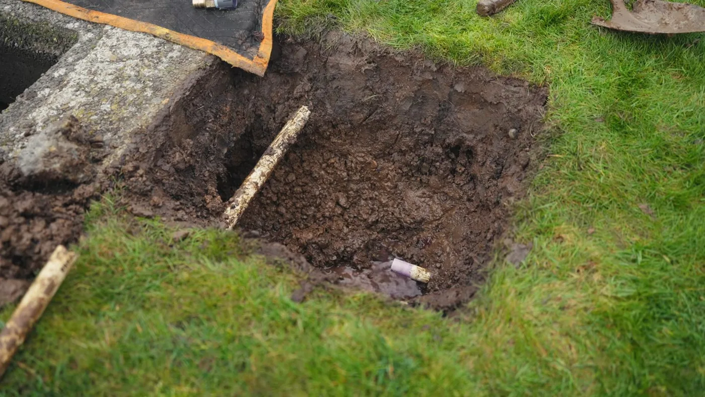 Square hole dug into a lawn beside a concrete slab, with loose soil and a short stick lying across it
