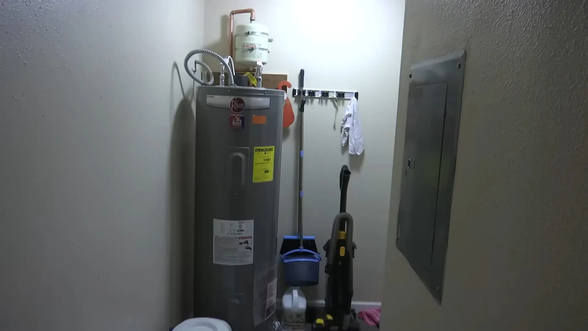 Electric tank water heater in a utility closet beside a mop, a vacuum and a wall panel