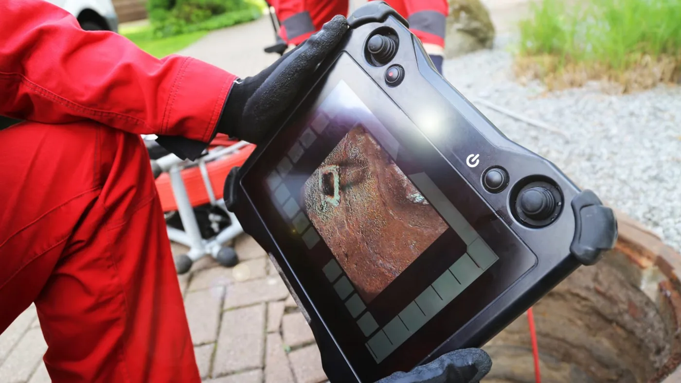 Technician in red coveralls holding a rugged camera monitor that shows the inside of a pipe