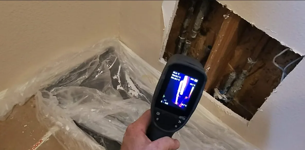 Hand holding a thermal imaging camera toward a cut opening in a wall, with a heat image on its screen