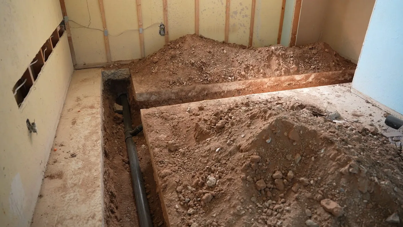 Trench cut into a basement floor beside a pile of loose soil in a framed room