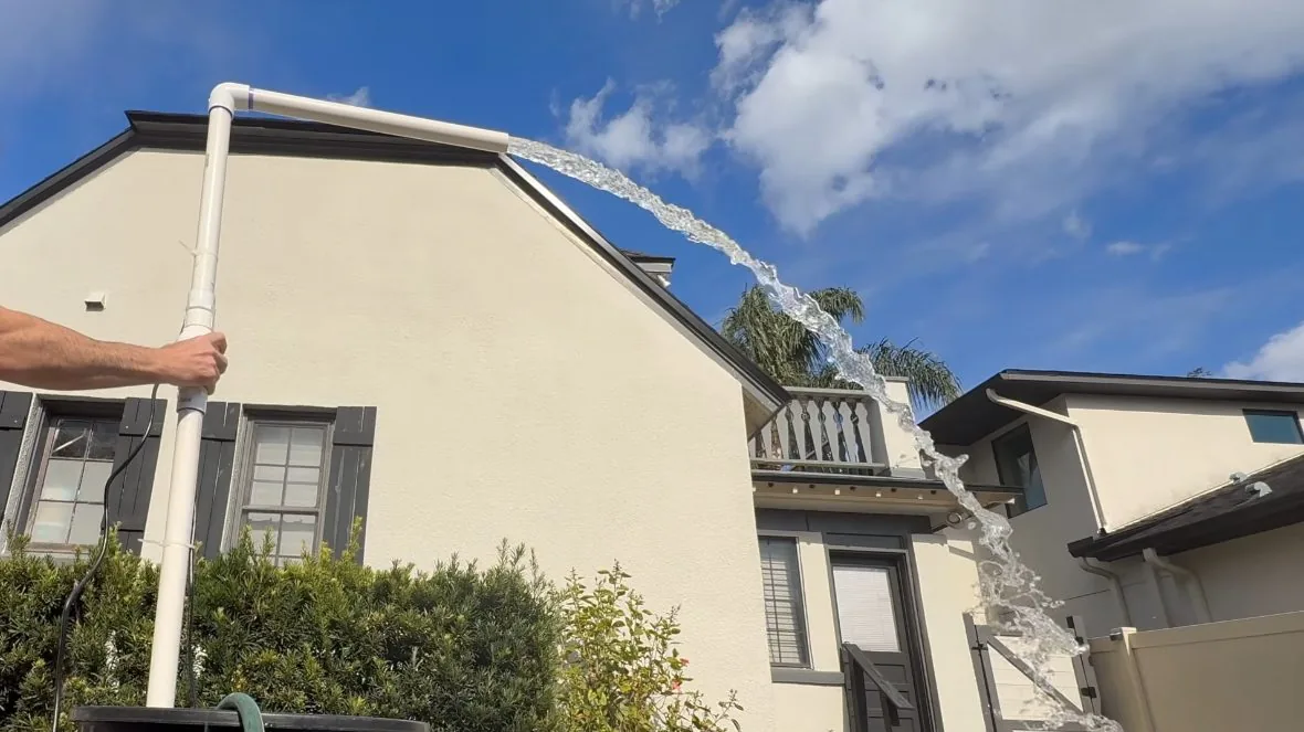 White pipe on the side of a stucco house with a stream of water arcing out of its top