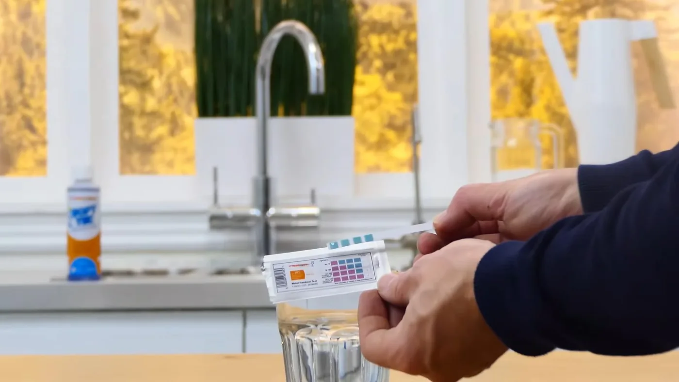 Hands holding a water test strip above a glass of tap water beside a kitchen faucet