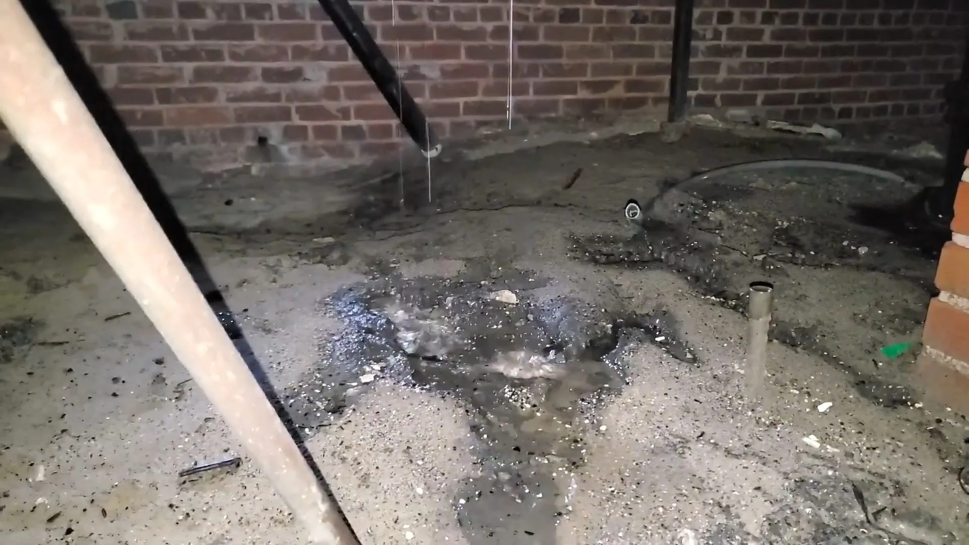 Basement floor covered with wet debris and standing water beside a brick wall, with a diagonal pipe at the left