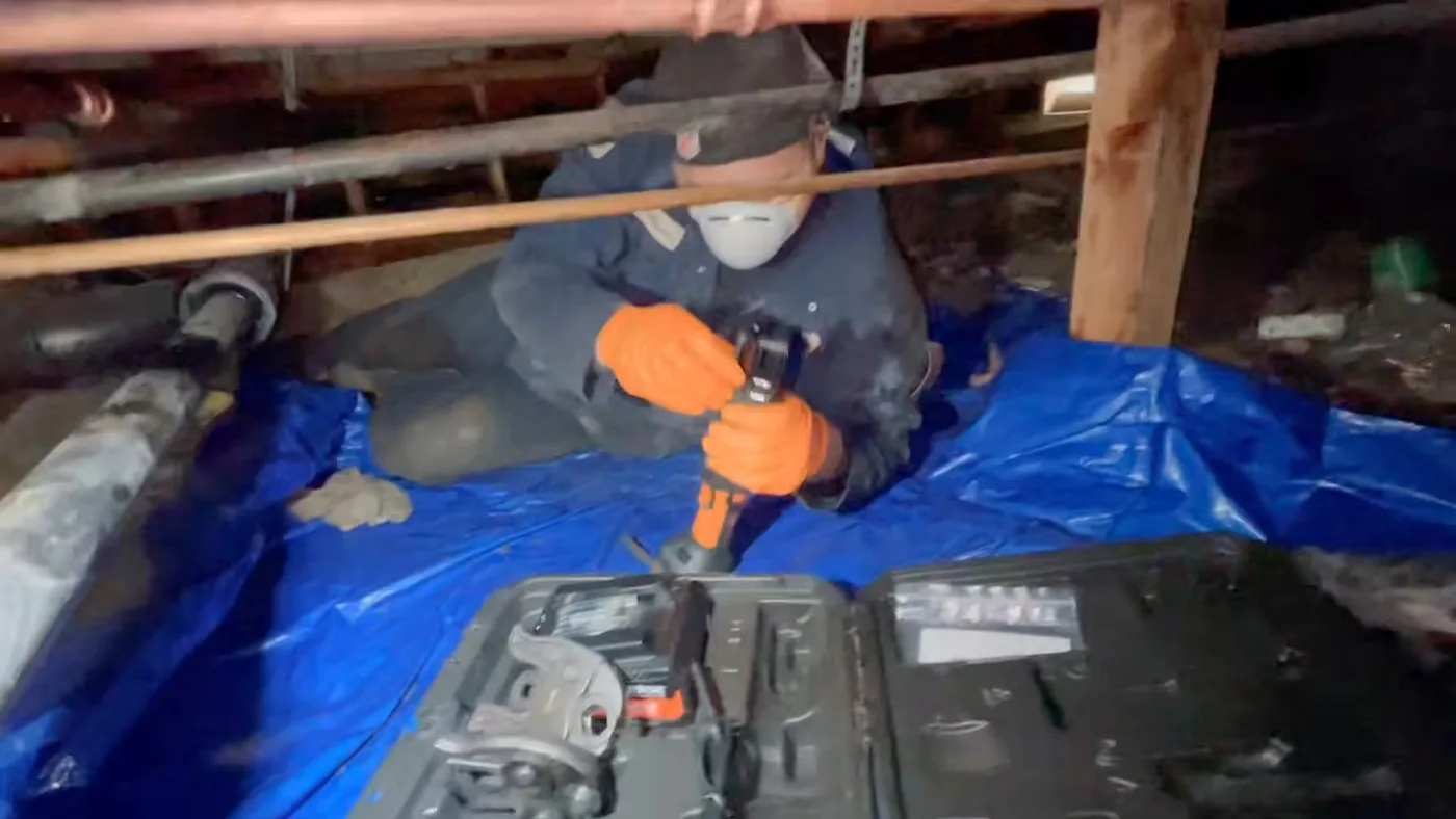 Worker in a mask and orange gloves kneeling on a blue tarp in a crawl space with a power tool in hand
