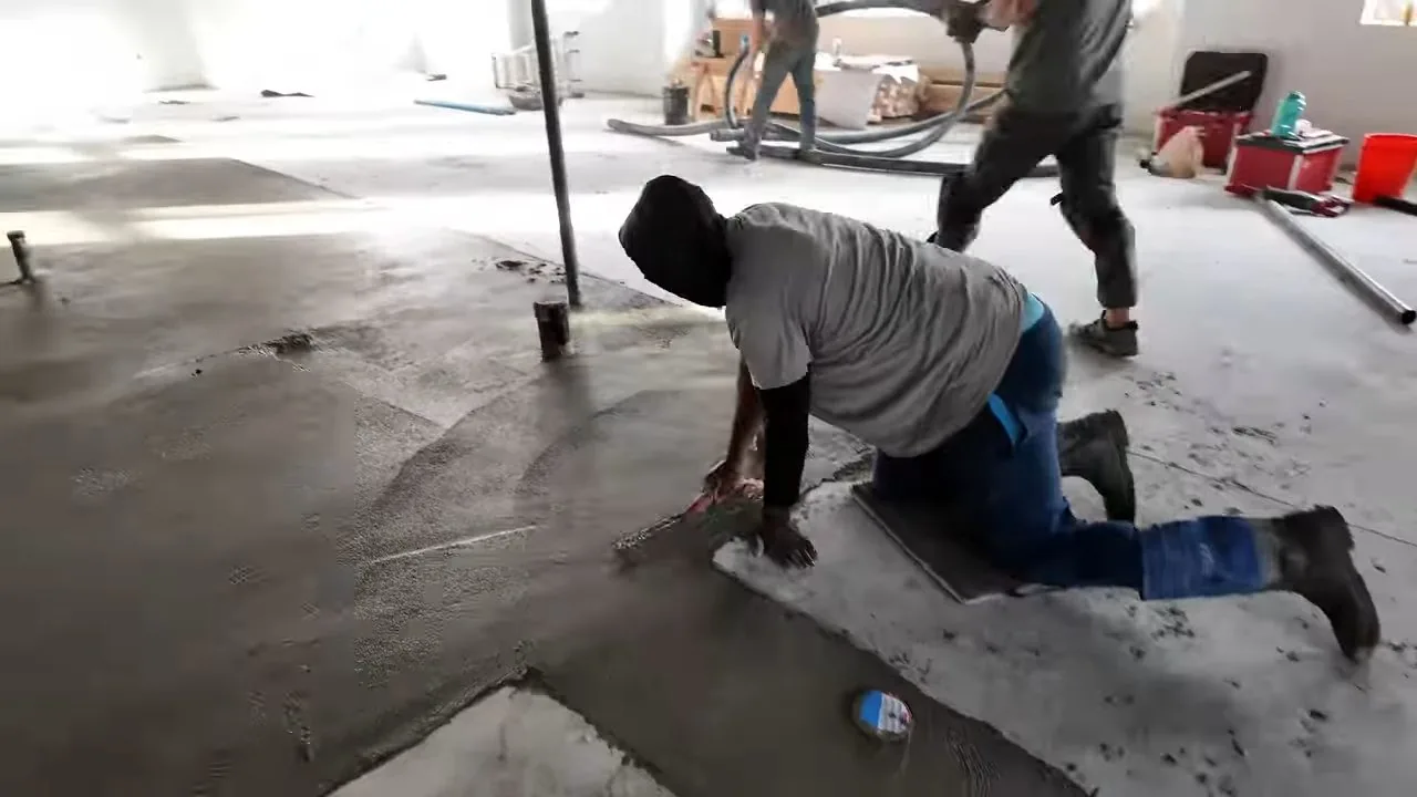 Worker kneeling on a bare concrete slab inside a building while another man works behind him