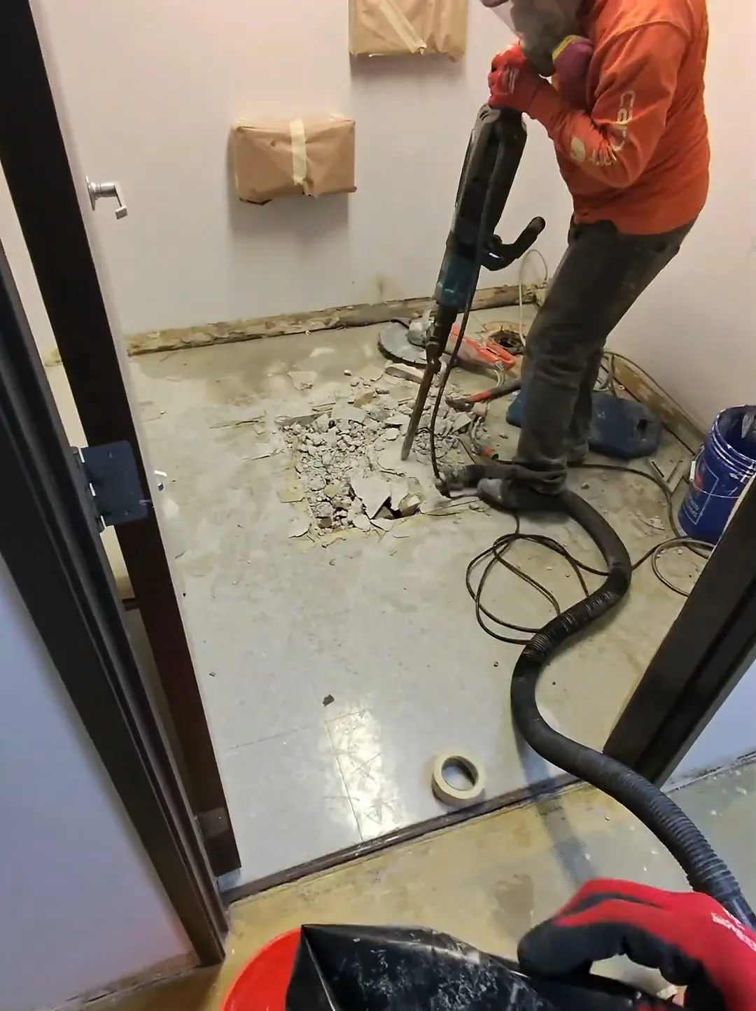 Worker in an orange sweatshirt using a jackhammer on a concrete floor in a small room